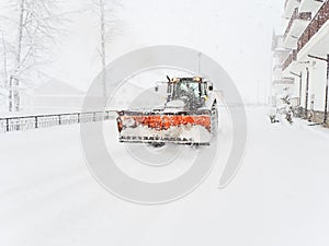 A snow plow tractor drives on a snowy road along a building during a severe blizzard