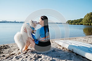 Snow-White Japanese Spitz Kissing Woman with Locs, Female Dog Owner Sitting on the Beach of City Lake