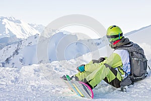 Snowboarder freerider sits on a slope and fastens his snowboard while looking at the mountains. Russia Sochi Rosa Khutor