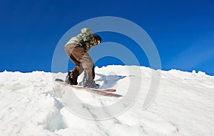 Snowboarder riding snowboard in deep snow down mountain slope on background of bright blue sky.