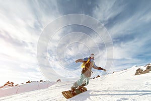 A snowboarder in a ski mask and a backpack is riding on a snow-covered slope leaving behind a snow powder against the