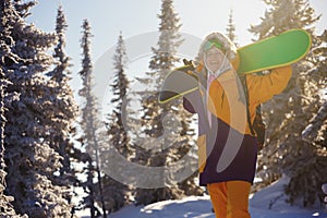 A snowboarder with a snowboard on her shoulders in the winter forest, the sun is shining from behind