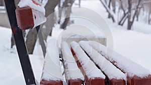 Snowfall in winter park, empty bench under snow with snowdrifts by walkway in alley at snowy season