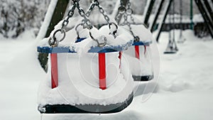 Snowflakes falling on empty swings at public playground and covering park with snow. Concept of winter holidays, bad weather and