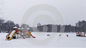 It is snowing in the city. A snowstorm is covering a playground with play structures on the edge of the lawn and surrounded by tre