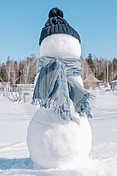 A snowman in a blue hat and scarf is standing in the field. An iceman on a winter forest background. Sunny day, freezing cold,