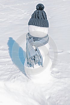 A snowman in a blue hat and scarf is standing in the white field. An iceman on a light showy background. Sunny day, freezing cold