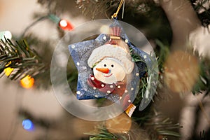 Snowman CHristmas tree ornament on green branch with string lights and copy space. Winter holidays background.