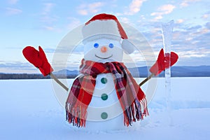 The snowman in plaid scarf, red hat, gloves. Amazing sunrise enlighten the sky. Nice landscape with the mountains. Winter day.