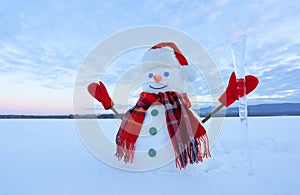 The snowman in plaid scarf, red hat, gloves. Amazing sunrise enlighten the sky. Nice landscape with the mountains. Winter day.