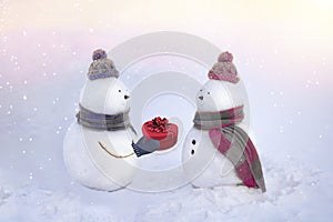 Snowmen in love wearing winter hats, scarves on a white background in pink and blue colors. Boy giving a gift to his