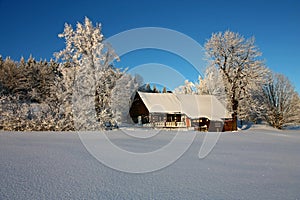 A traditional Czech cottage is nestled in a pristine, snow-covered winter landscape under a clear blue sky.