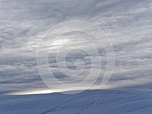 Snowy hill by rippled clouds at sunset, pale light in winter
