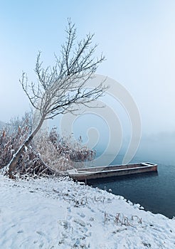 Snowy landscape of frozen lake in winter with fishing boat ashore under frozen tree