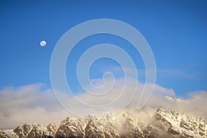 A snowy mountain range with a full moon in the clear dip blue sky.