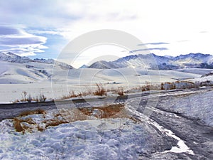 Snowy mountains and ridge  country road with puddle  brown grass in the foreground. Winter landscape