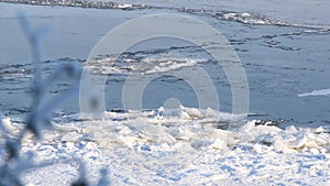 A snowy river bank with ice floes floating along the river, and blurred bush branch in foreground