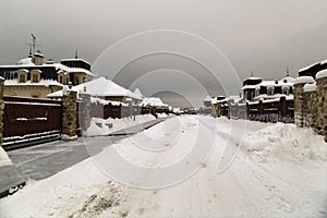 Snowy road between country houses. Winter landscape with snowy street, brick sidewalk, cottage houses and gray sky.