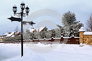 Snowy road between country houses. Winter landscape with street lamp, street signs, sidewalk, snow footprints. Street landscape