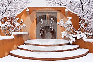 Snowy scene of adobe wall with rustic wood doors and chile ristras