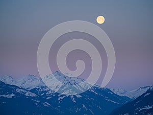 Snowy winter alpine landscape after sunset, pink purple sky with a full moon over a mountain peak