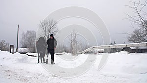 snowy winter, disabled man jockey leads, holding with reins a black horse on the way. man has a prosthesis instead of