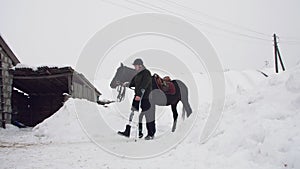 snowy winter, disabled man jockey leads, holding with reins a black horse on the way. man has a prosthesis instead of