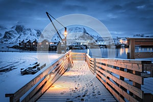 Snowy wooden pier on the sea coast with lights, rorbu, mountains