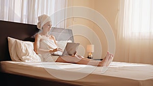 While snuggled up in a cozy bathrobe and king size bed, a young woman typing on her laptop proves she can work from