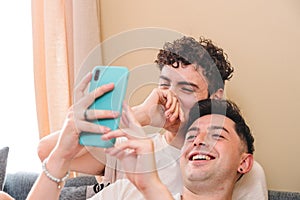 Snuggling gay couple in pajama laughing and using the smart phone. Cohabitation of homosexual couple