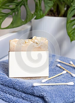 Soap bar with blank label on blue towels near basin and green monstera in bath, mockup
