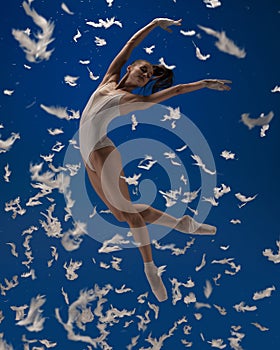 Soar like a bird. One young female ballet dancer flying isolated over blue background with white feathers.