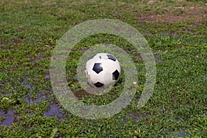 A soccer ball lies on the grass among puddles and ridges in summer. There is a ball in a puddle on the football field