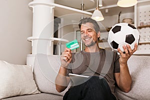 Soccer fan man watching football game on television at living room couch. He holds a credit card and a digital tablet for