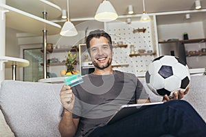 Soccer fan man watching football game on television at living room couch. He holds a credit card and a digital tablet for