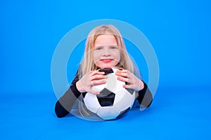 Soccer fans. Cute blonde girl in black shirt holding soccer ball in hands over blue studio background. Copyspace