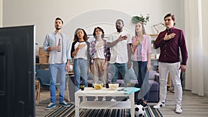 Soccer fans singing anthem with hand on chest standing at home watching TV