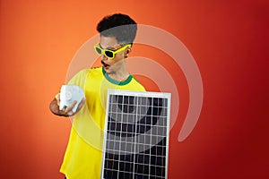Soccer Player - Black Man Celebrating Holding Solar Photovoltaic Panel and Pig Coin Isolated on Orange Background
