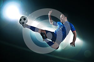 Soccer player in mid air kicking the soccer ball, stadium lights at night in background