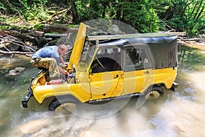 Off-road Russian car stuck in mountain river at sudden breakdown while jeeping. Driver fixing the auto with hood lifted up