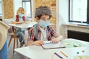 Social distancing. School boy wearing face mask writing in his notebook while sitting at the desk in a classroom. Kids