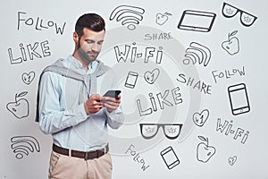 Social media concept. Handsome young man in casual wear using his smart phone while standing against grey background