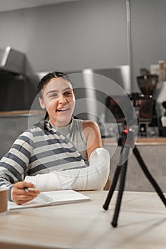 Social media influencer or student blogger creating content in cafe. Young woman with broken hand using smartphone on