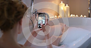 Social media, phone and woman relax in bath with bubbles, candles and post self care in home. Online, photography and