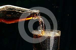 Soda, coke, dark beer pouring from bottle into glass against black background