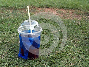 Close-up soda pop in transparent plastic cup with white straw and blue bag on green grass field
