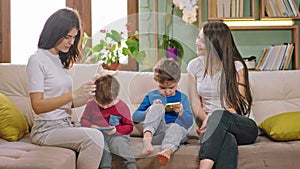 On the sofa two ladies young mothers with their kids two pretty boys chatting and smiling while the kids are playing a