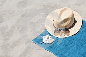 Soft blue towel, sunglasses, straw hat and seashell on sandy beach, space for text