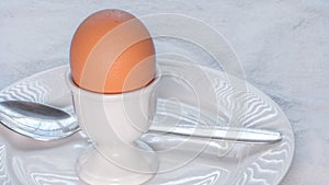 Soft boiled brown egg in a white ceramic egg cup, on a white ceramic plate with a spoon, on a white cloth tablecloth