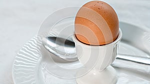 Soft boiled brown egg in a white ceramic egg cup, on a white ceramic plate with a spoon, on a white cloth tablecloth